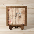 May include: A wooden framed sign with the word "joy" in white script. The sign features a floral and script design on a wooden background. The sign is displayed on a wooden easel.