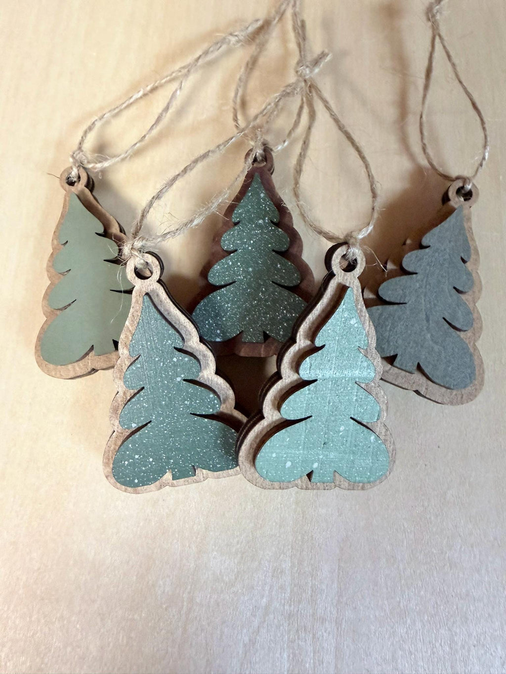 May include: A set of five wooden Christmas tree ornaments in various shades of green. Each ornament features a layered design with a natural wood base and a painted top layer, some with a speckled finish. Each piece includes a thin jute twine loop.