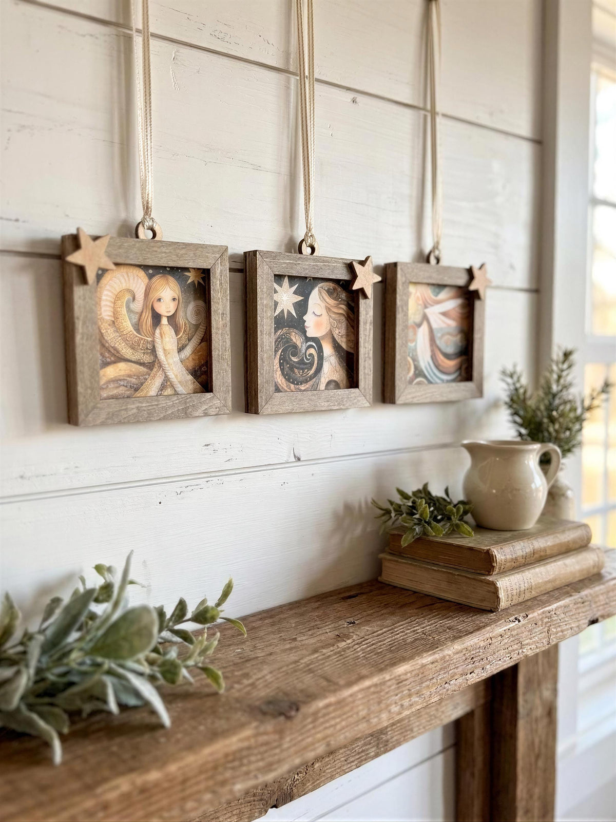 a rustic wooden shelf with three framed pictures hanging on it, each depicting a different scene. The shelf also holds a small white pitcher, a stack of books, and some greenery, creating a cozy and inviting atmosphere.