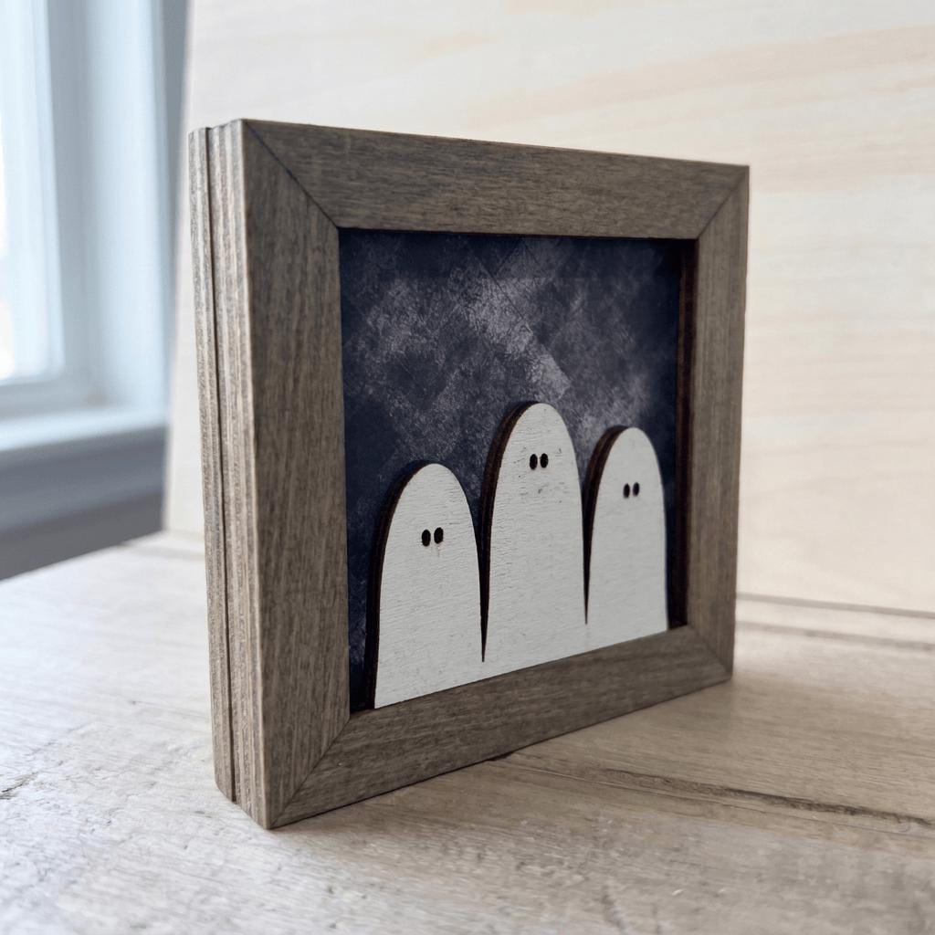 A wooden frame containing a framed image of three white ghost figures against a dark background.